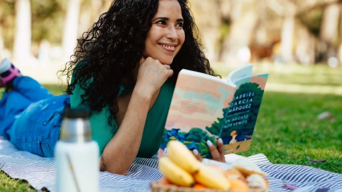 Smiling woman reading a book at a picnic in a sunny park setting.